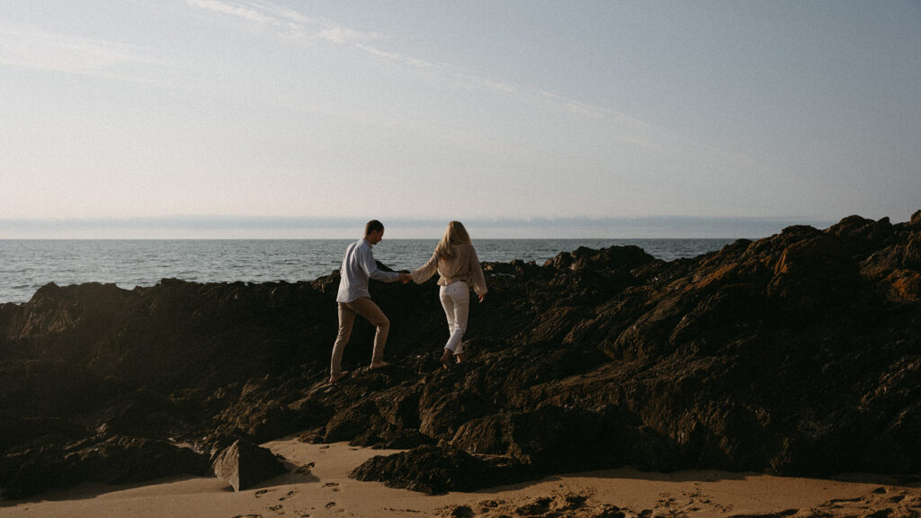 Elodie et Gurvan grimpant sur des rochers à la plage pendant une séance photo d'engagement.