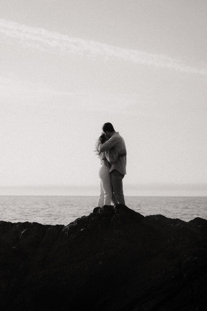 Elodie et Gurvan enlacés sur les rochers face à la mer. Photo en noir et blanc.